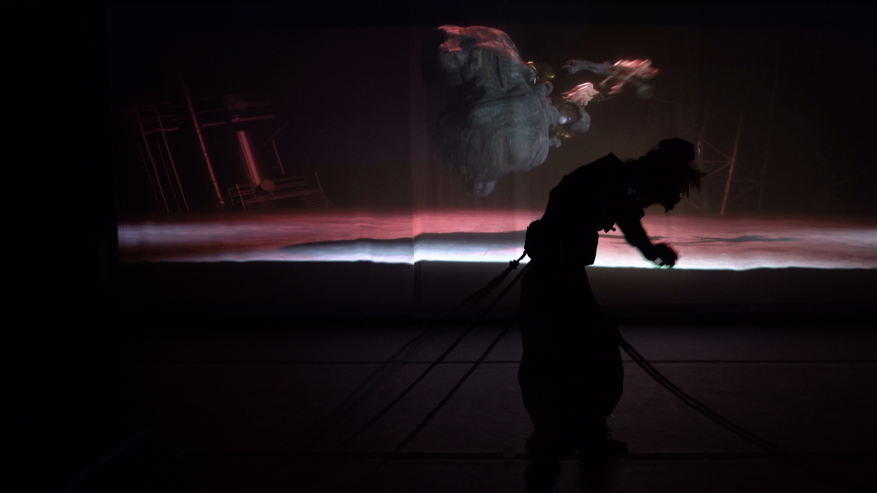 Silhouetted figure bending forward with several ropes or cords attached, against a dark background with a faint abstract projection in shades of red and white.