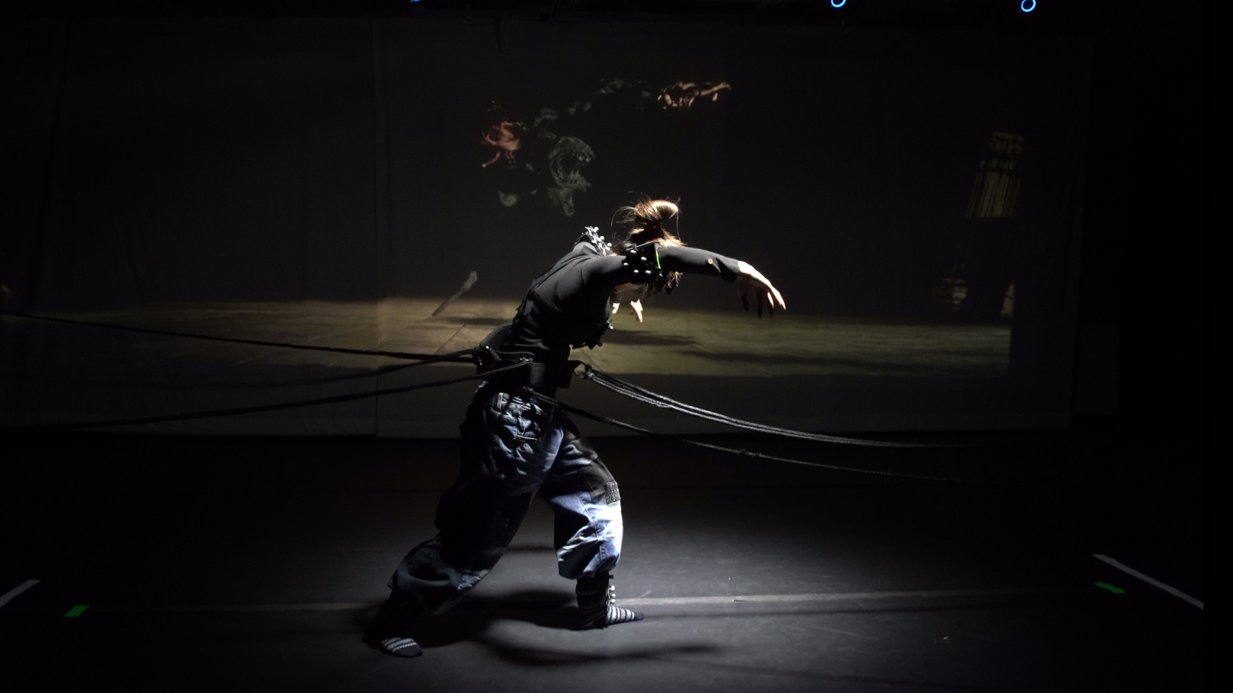 Person in dark clothing striking a dynamic pose with arms extended, tethered by multiple ropes in a dimly lit space with a projection in the background.
