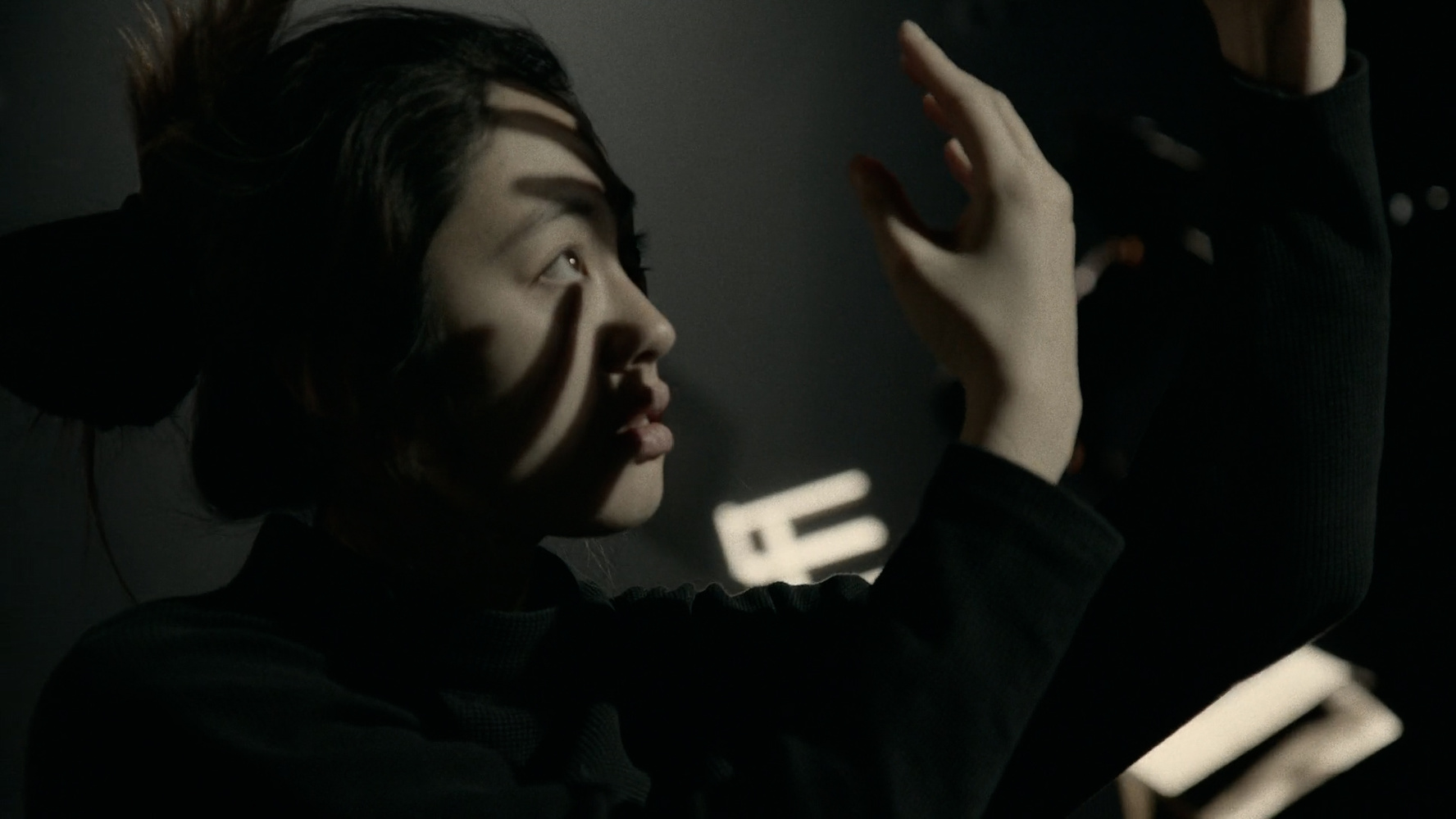 Woman with hair in a bun looking upward with hands raised, shadows cast on her face in a dark setting.