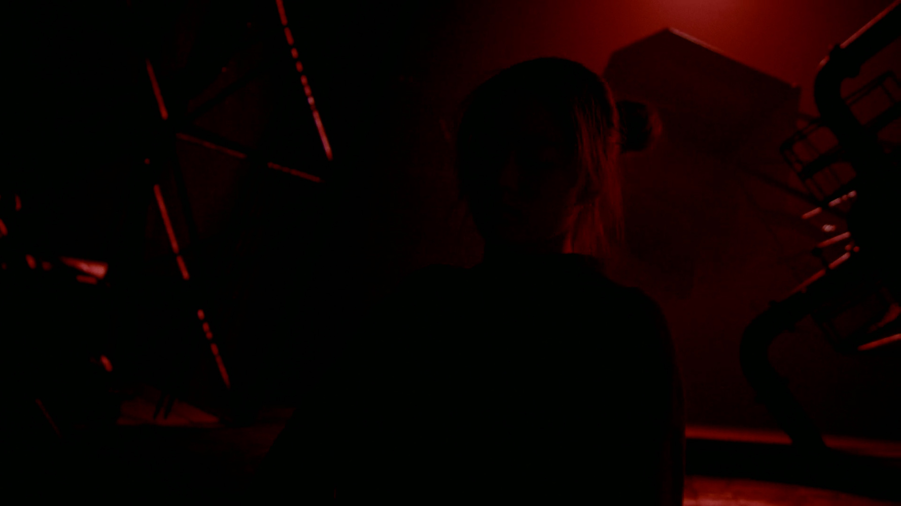 Silhouette of a person with hair tied back illuminated by red light in a dark industrial setting.